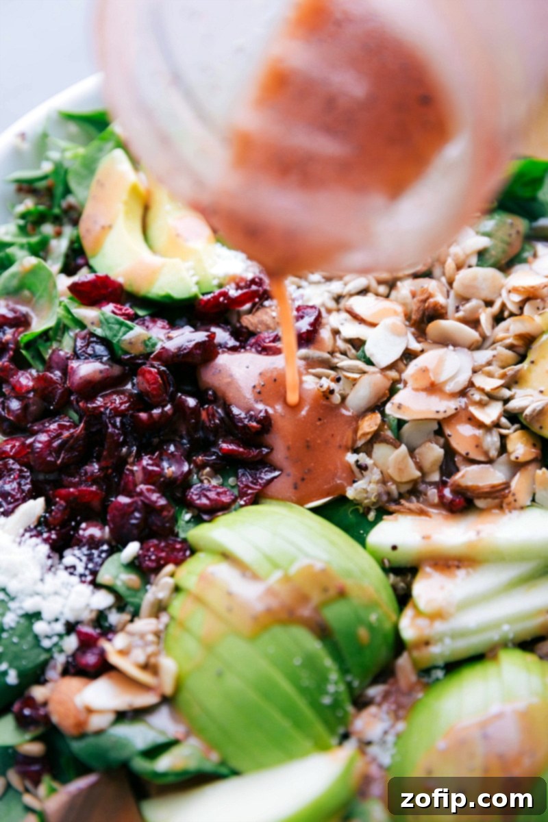 A hand drizzling generous amounts of raspberry vinaigrette over a large bowl of Quinoa and Almond Salad, showcasing the rich colors of the ingredients.