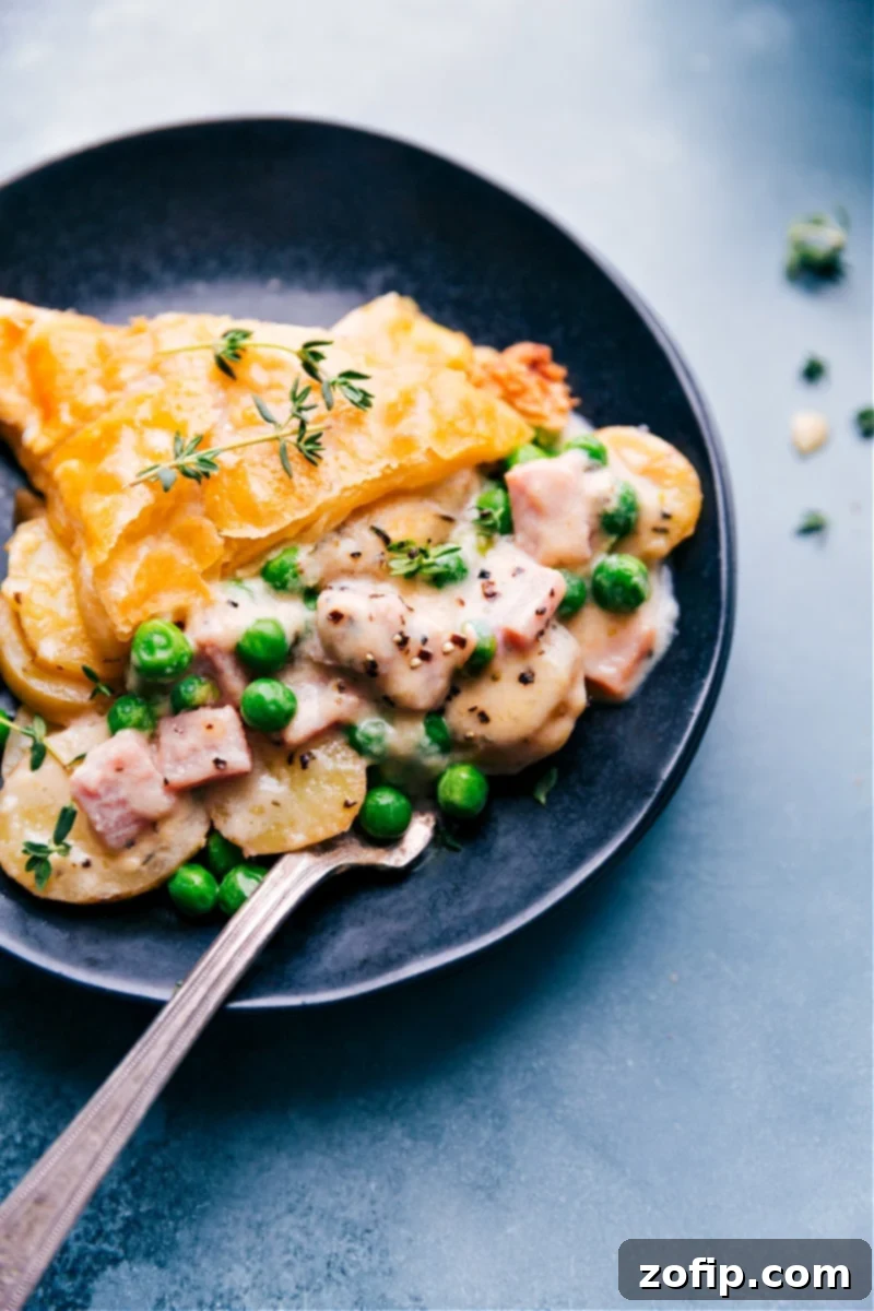 Delicious Ham and Potato Pot Pie Slice On A Plate A generous slice of Ham and Potato Pot Pie on a rustic plate, revealing layers of cheesy potatoes, ham, and peas beneath a golden, flaky puff pastry crust.