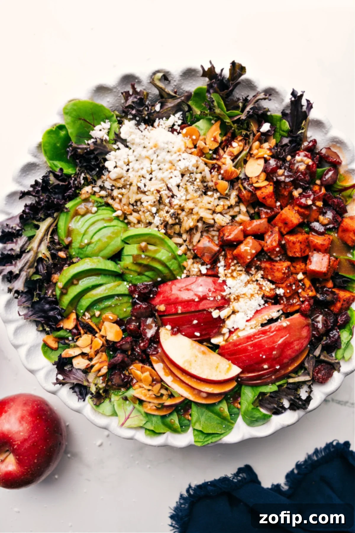 A vibrant Fall Salad in a large wooden bowl, artfully arranged with roasted sweet potatoes, mixed greens, apple slices, cranberries, and almonds, with a drizzle of lemon balsamic dressing.