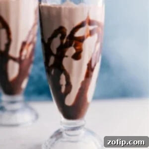 A tall glass of frothy Malted Milkshake, topped with whipped cream and a cherry, against a blurred background.