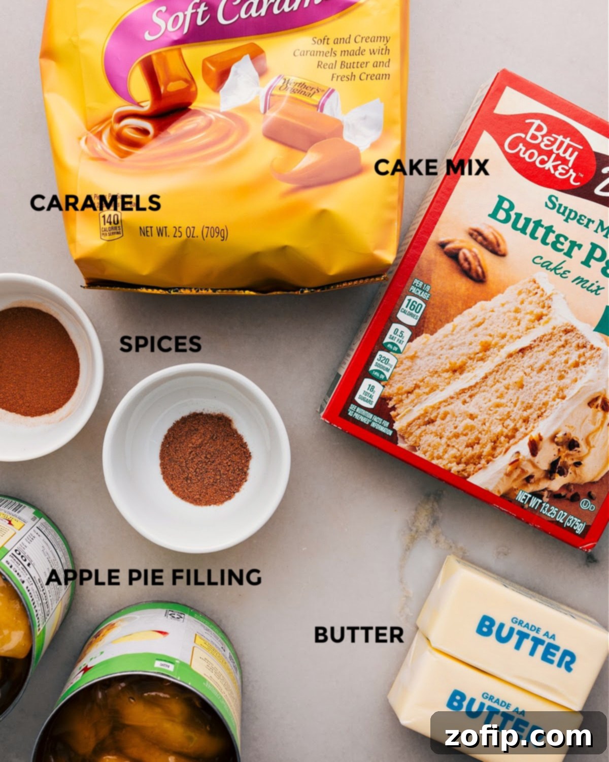 All the essential ingredients for Apple Dump Cake laid out: cans of apple pie filling, butter pecan cake mix, ground cinnamon, unsalted butter sticks, and a bag of soft caramels.