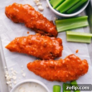 Close-up of baked Buffalo Chicken Tenders coated in sauce, with a few extra tenders on the side and a dipping bowl.