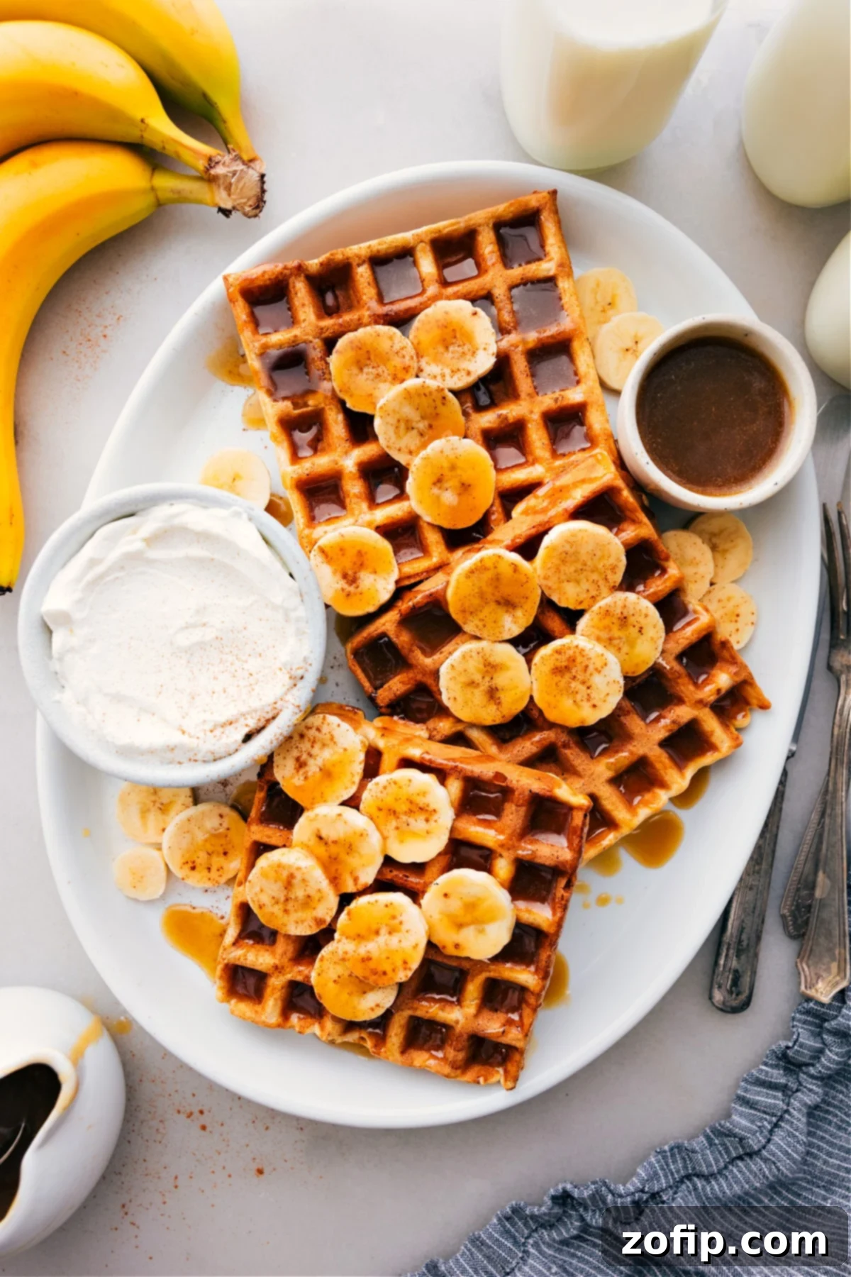 A beautiful platter of golden-brown banana waffles, generously drizzled with rich maple syrup and garnished with fresh banana slices and a dollop of creamy whipped cream. A perfect, inviting breakfast spread.