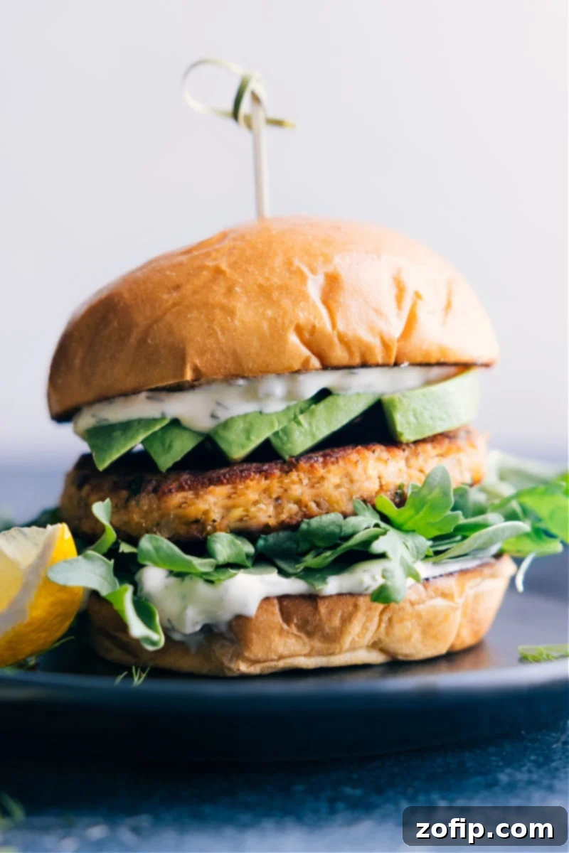 A succulent Salmon Burger on a brioche bun, complete with fresh greens, creamy avocado, and a generous dollop of homemade lemon-dill tartar sauce, served on a white plate.