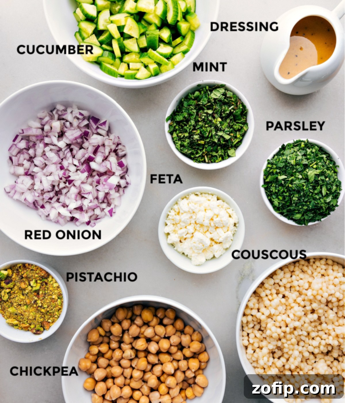 All the fresh ingredients for the Jennifer Aniston Salad are prepped and laid out for easy assembly, including vibrant cucumbers, fresh mint, parsley, creamy feta, hearty couscous, crunchy pistachios, sharp red onion, and protein-rich chickpeas, with the dressing on the side.