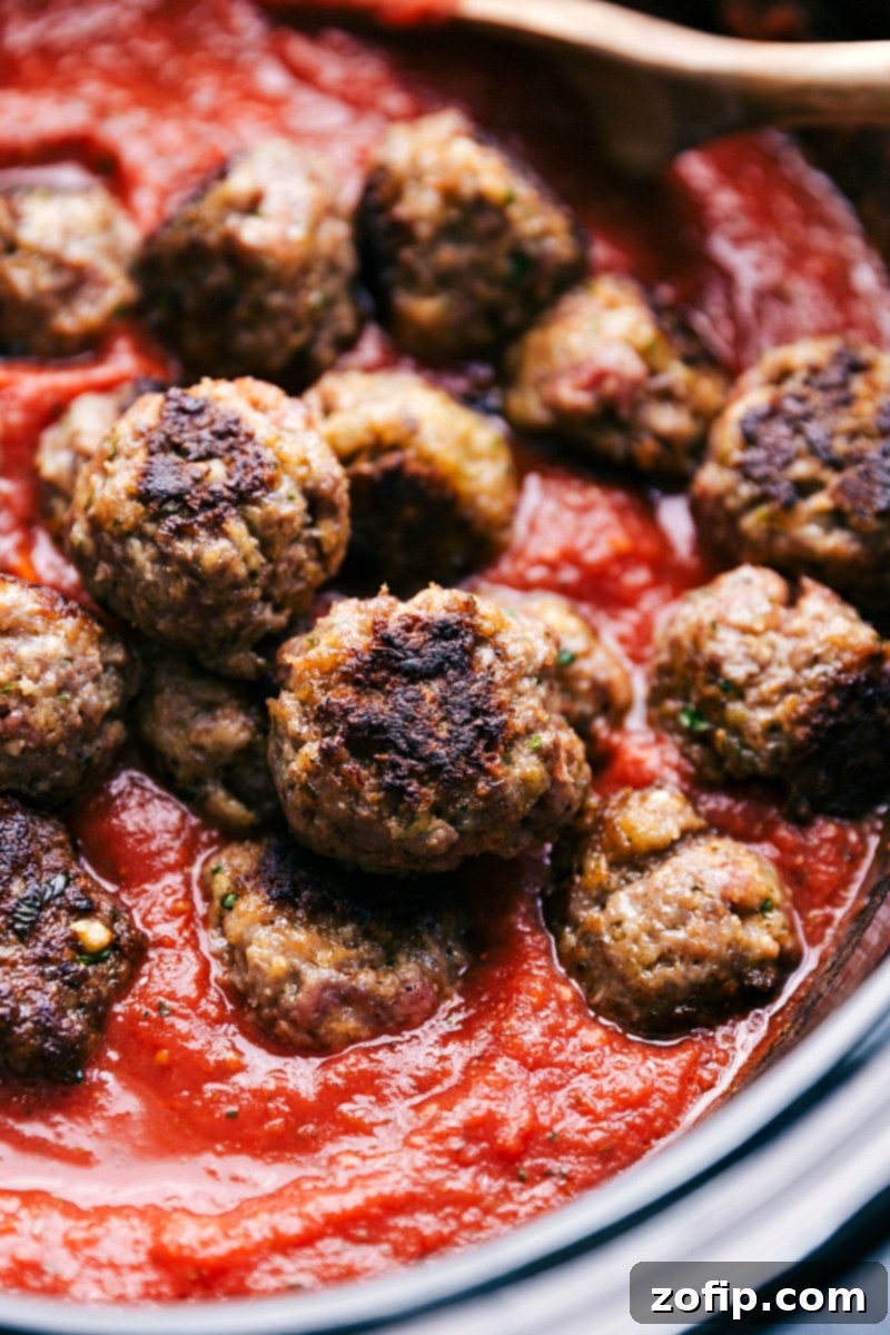 Tender and flavorful meatball sliders with an incredible homemade marinara sauce made in the slow cooker. Add the meatballs and sauce to a slider roll, add cheese, and broil them to make the ultimate meatball sliders. Close-up image of homemade meatballs being gently placed into rich marinara sauce, ready for slow cooking