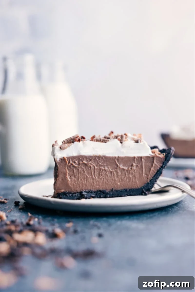 A perfectly sliced piece of Chocolate Cream Pie on a white plate, showcasing its layered texture with a dark crust, rich chocolate filling, and white whipped cream topping, garnished with chocolate shavings.