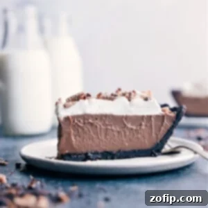 A slice of rich Chocolate Cream Pie on a white plate, showcasing its layers of dark Oreo crust, smooth chocolate pudding, and fluffy whipped cream, topped with chocolate shavings.