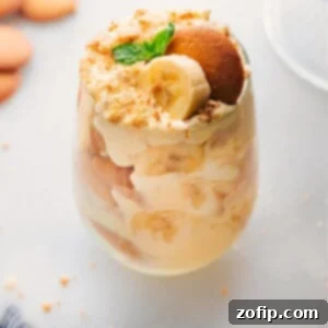 A glass filled with layers of the delicious banana pudding recipe, alternating between pudding, vanilla cookies, fresh bananas, and topped with vanilla cookie crumbs, creating a delightful dessert.