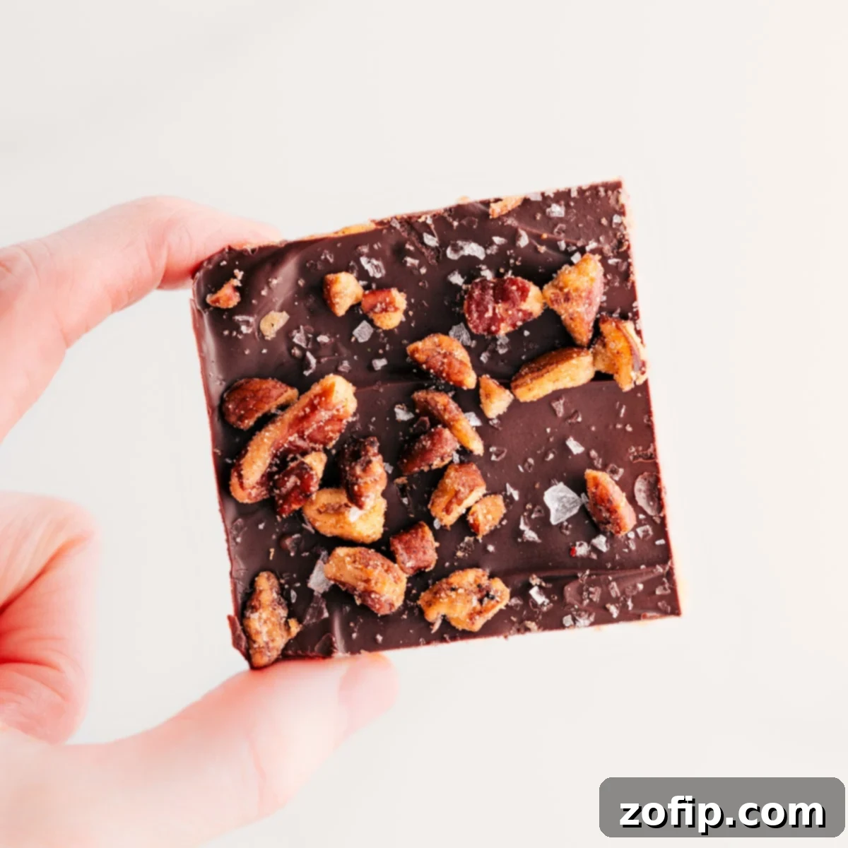 Christmas Crack Topped With Candied Pecans A single square of Christmas Crack is held up, showcasing its decadent layers of salty cracker, golden toffee, rich chocolate, and a generous topping of candied pecans.