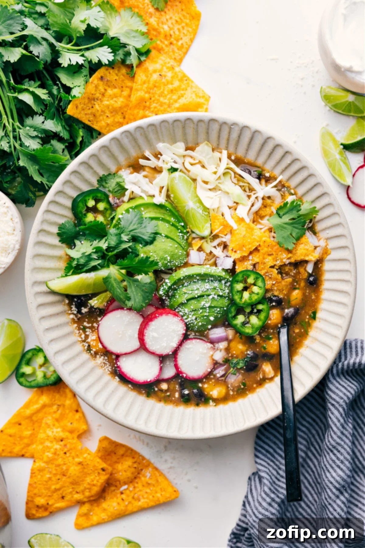 Chicken Pozole served in a bowl topped with shredded cabbage, tortilla chips, avocado slices, radishes, cilantro, lime wedges, and jalapeño.