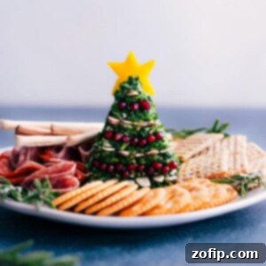 Beautifully decorated Christmas Tree cheeseball on a festive serving platter, adorned with sliced almonds, pomegranate arils, and a yellow bell pepper star, surrounded by crackers.
