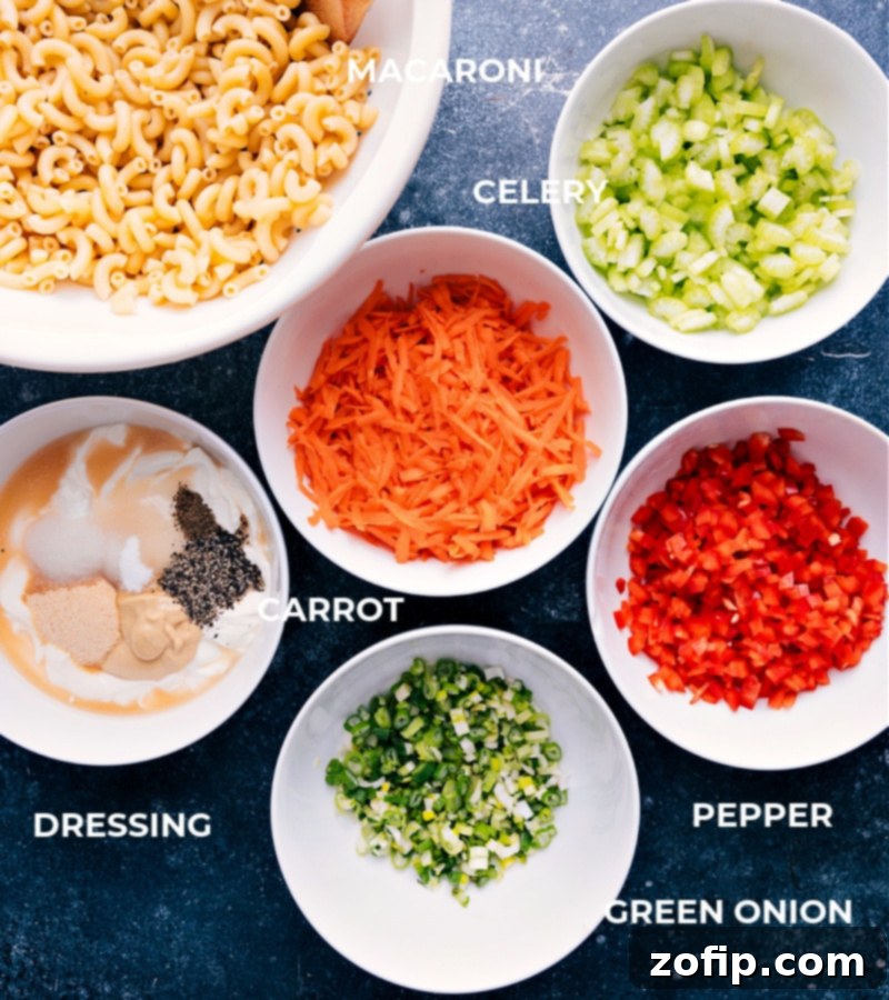 Various ingredients for Macaroni Salad laid out on a white surface, including cooked elbow macaroni, chopped celery, grated carrots, sliced green onions, red bell pepper, and separate bowls of mayonnaise, sour cream, and spices.