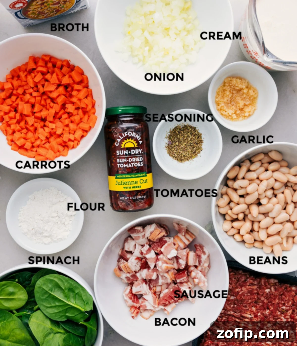 A flat lay photograph displaying all the fresh and essential ingredients for Tuscan Soup: vibrant vegetables, aromatic seasonings, sun-dried tomatoes, white beans, uncooked sausage, crispy bacon, fresh spinach, flour, chicken broth, and heavy cream.
