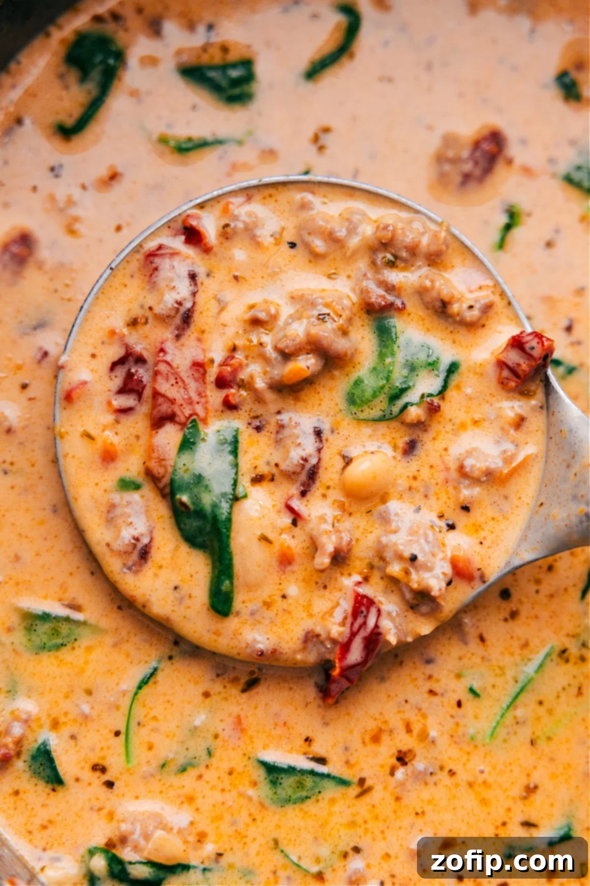A generous scoop of the finished Creamy Tuscan Soup, brimming with Italian sausage, white beans, spinach, and sun-dried tomatoes, being lifted from a large pot with a ladle.