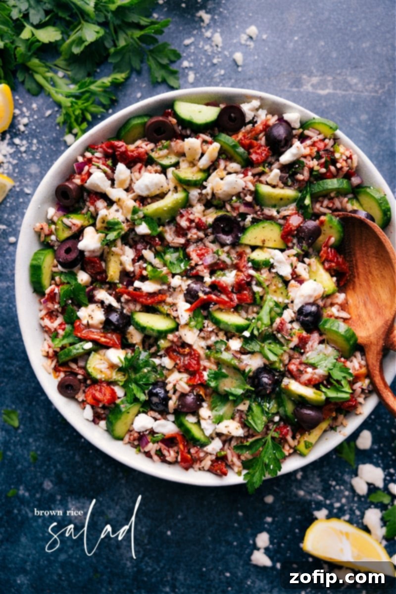 Overhead shot of a vibrant Brown Rice Salad in a large serving bowl, featuring nutty brown rice mixed with diced red peppers, cucumbers, sun-dried tomatoes, fresh parsley, and crumbled feta cheese, all glistening with a lemon dressing.