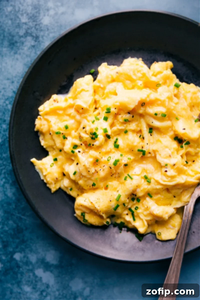 Close-up overhead image of a plate of perfectly cooked scrambled eggs, garnished with fresh green chives, showcasing their creamy, custardy texture and readiness for breakfast