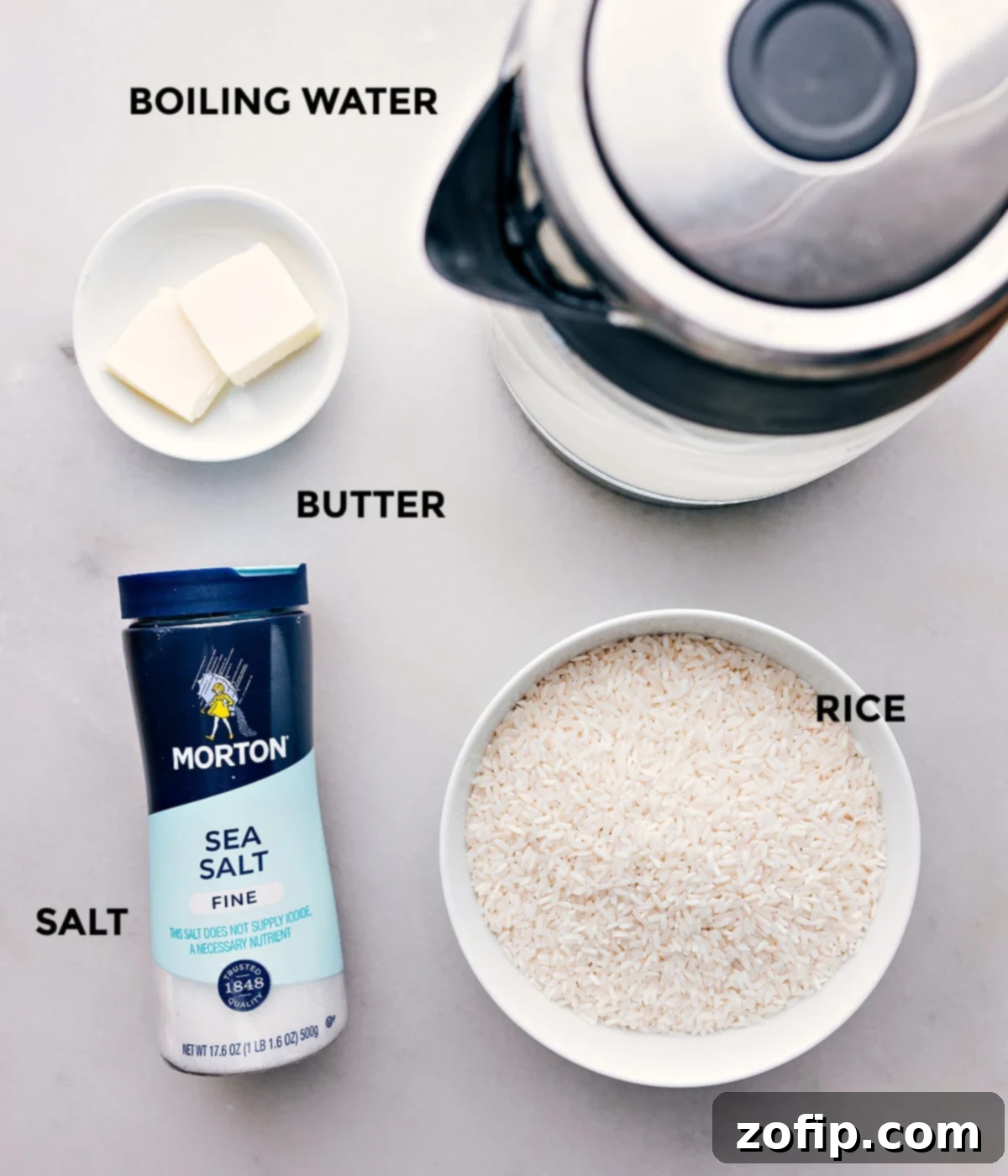 All the essential ingredients for oven-baked white rice laid out: rice, butter, salt, and a kettle of boiling water.