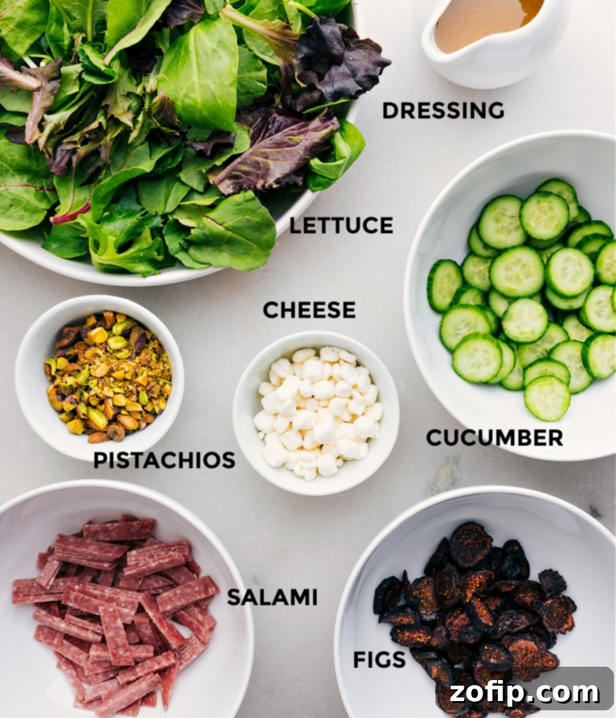 All the fresh ingredients for this Arugula Salad, including greens, dressing components, cucumbers, goat cheese, pistachios, salami, and dried figs, are neatly prepped for easy assembly.