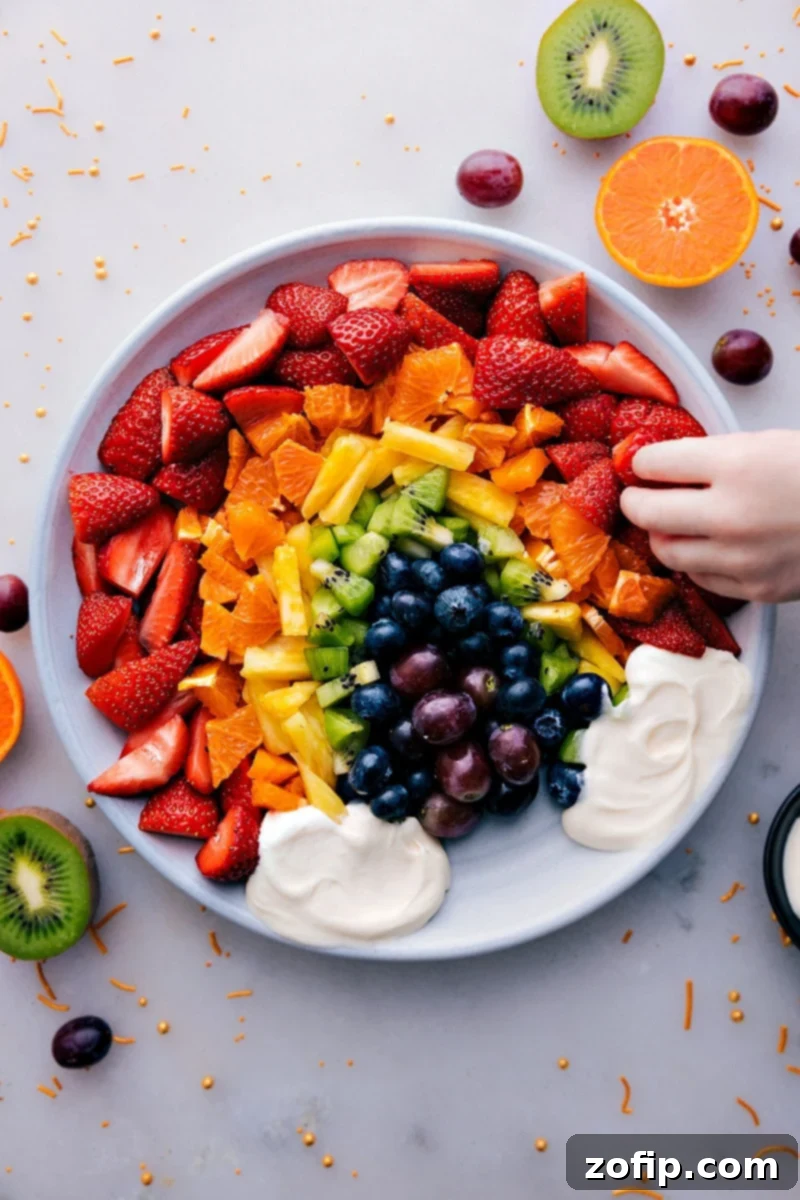 An overhead shot of a beautifully arranged Fruit Rainbow Tray with a cloud-like creamy dip at the bottom. The fruits are meticulously organized by color: red strawberries, orange mandarins, yellow pineapple, green kiwi and grapes, and blue/purple blueberries and blackberries, creating a stunning edible rainbow.