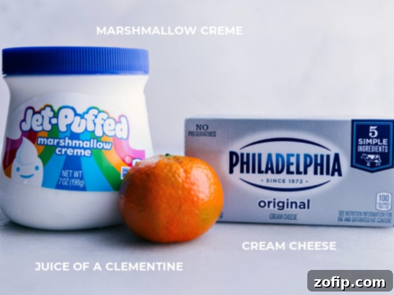 A close-up image showing the three key ingredients for the creamy fruit dip: a solid block of full-fat cream cheese, a transparent container of fluffy marshmallow creme, and a fresh clementine orange, all arranged neatly on a surface.