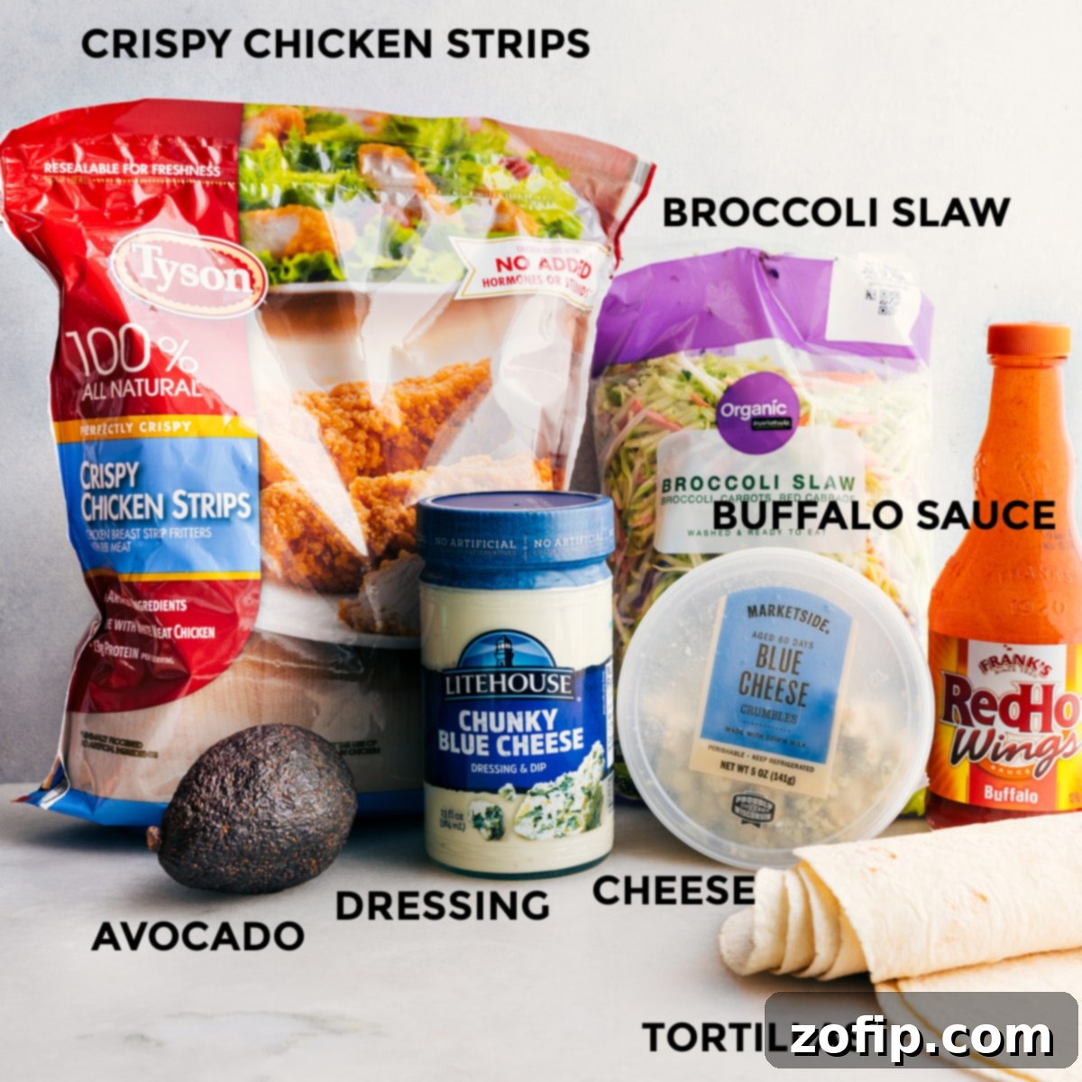 All the fresh ingredients for Buffalo Chicken Wraps laid out on a cutting board: crispy chicken strips, buffalo sauce, blue cheese dressing, broccoli slaw, tortillas, cheese crumbles, and sliced avocado.