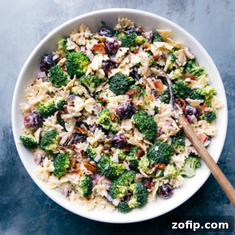 Broccoli Grape Pasta Salad, Perfectly Presented and Ready to Enjoy Overhead view of a beautifully prepared Broccoli Grape Pasta Salad presented in a clean white bowl, garnished with a sprig of fresh herbs, indicating it's perfectly ready for serving. The vibrant colors of green broccoli, red grapes, and light pasta are prominent.