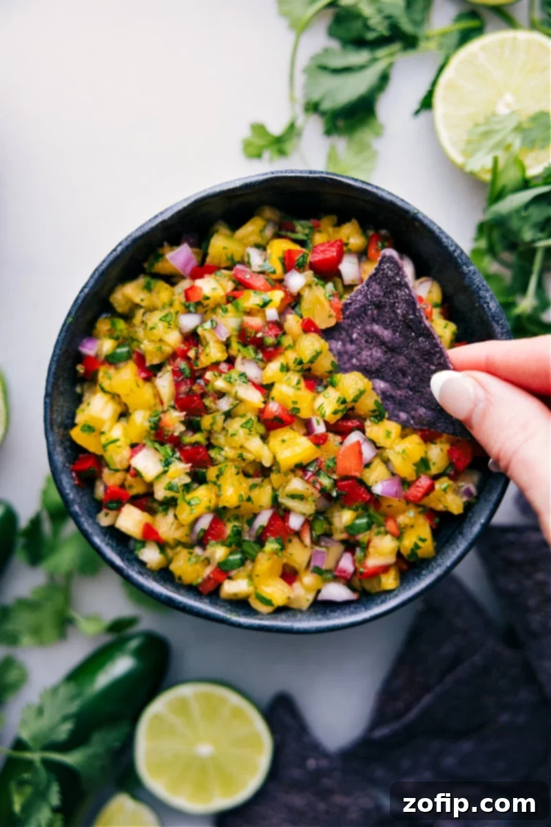 A vibrant bowl of fresh pineapple salsa, with a tortilla chip dipping in, showcasing its juicy chunks of pineapple, red onion, red pepper, and cilantro.