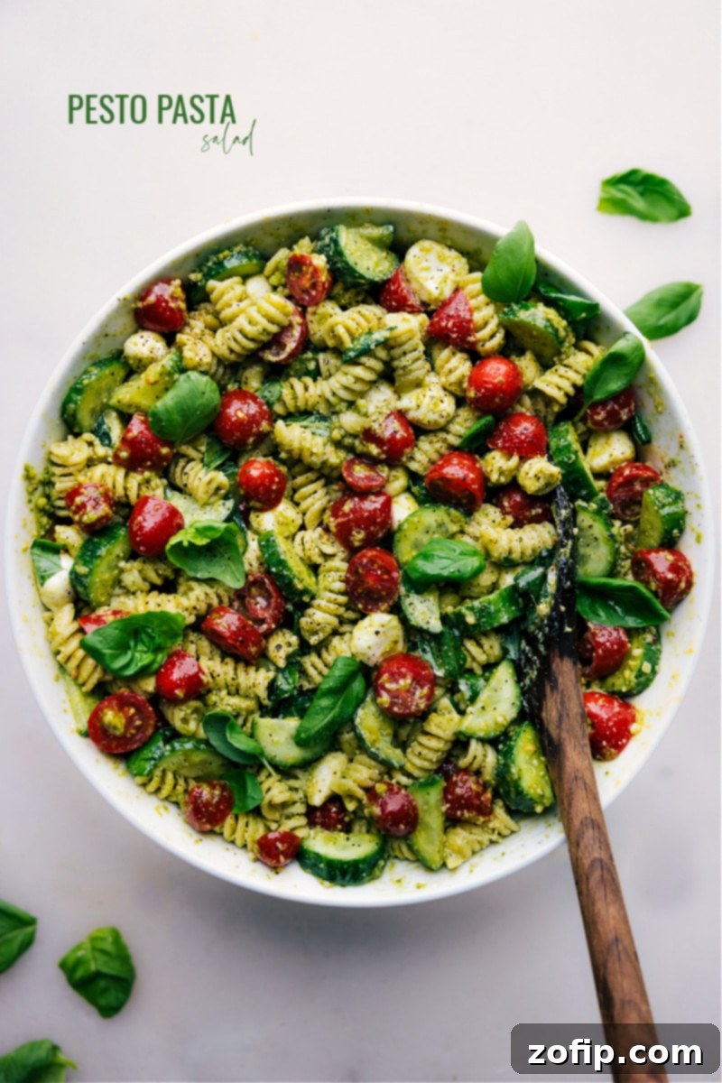 Overhead image of the vibrant Pesto Pasta Salad, showcasing rotini pasta, halved cherry tomatoes, sliced cucumbers, and mozzarella pearls, all coated in a rich green pesto sauce.