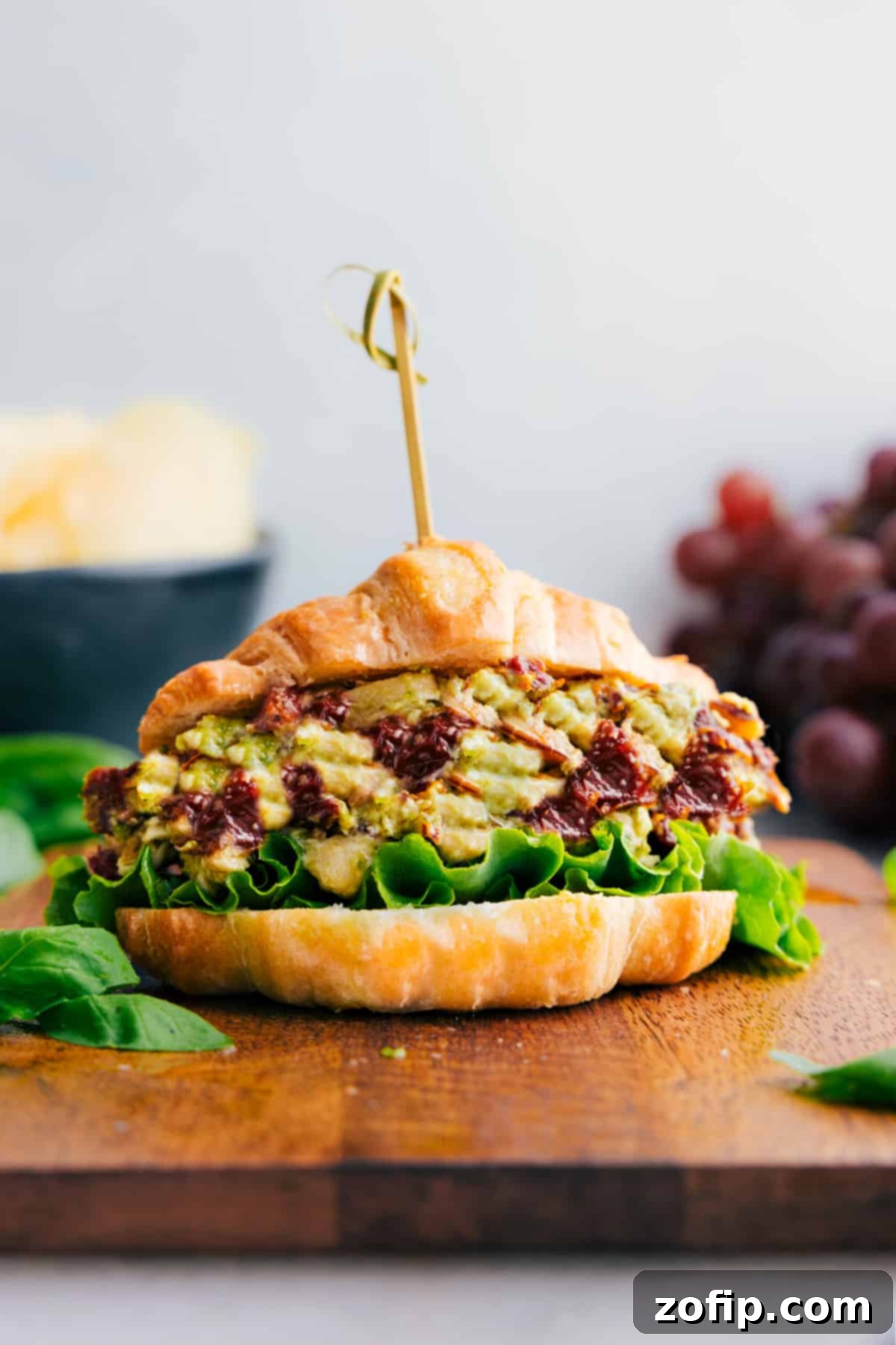 A perfectly assembled sandwich featuring Pesto Chicken Salad, served alongside fresh grapes and crunchy chips, ready for a delightful and complete meal.