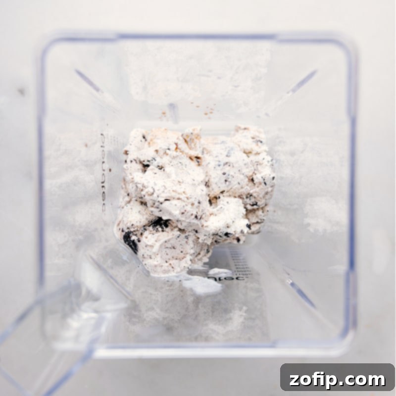 Starting the blending process: ice cream and milk go into the blender for our Oreo Milkshake. A scoop of cookies and cream ice cream being carefully added into a high-powered blender, ready to be transformed into a delectable Oreo Milkshake.