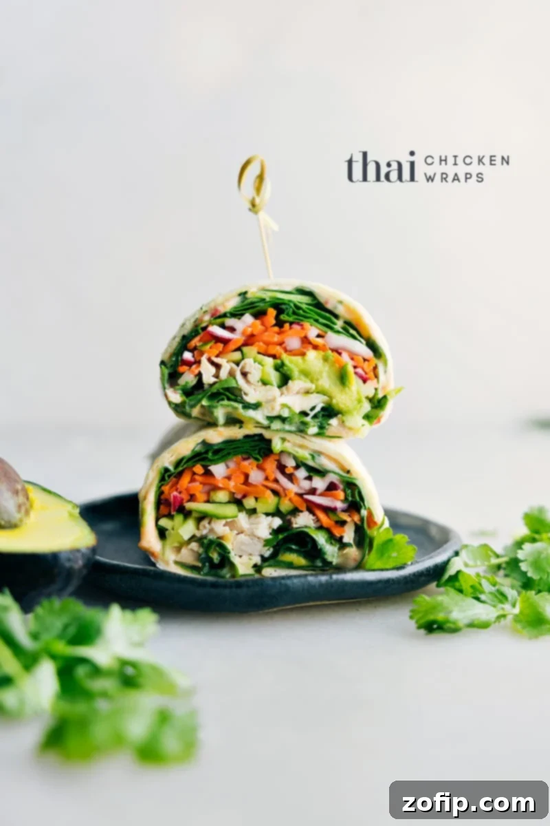 Two Thai Chicken Wraps on a rustic plate, garnished with fresh cilantro, ready to be enjoyed. The wraps reveal their colorful fillings of shredded chicken, vibrant pickled carrots and radishes, green spinach, and creamy avocado, making them look incredibly appealing and fresh.