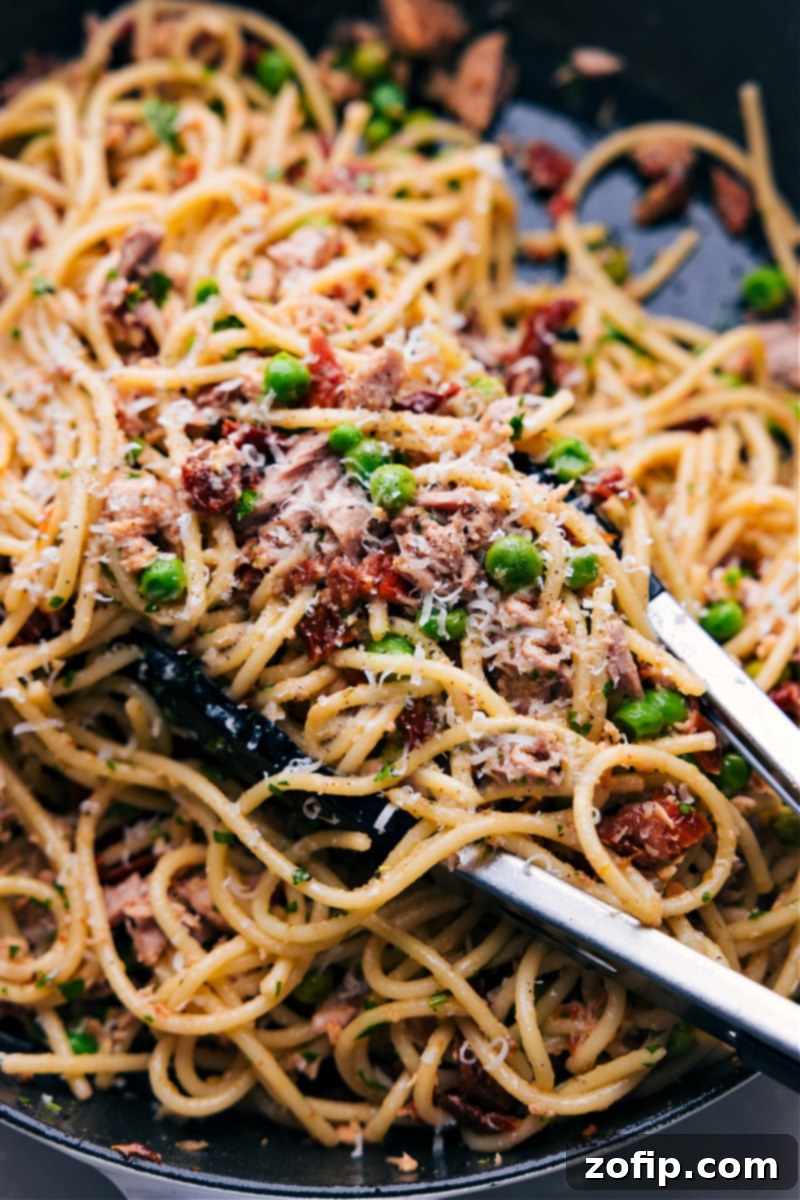 Serving Healthy Tuna Pasta with Tongs Tongs gracefully lifting a generous serving of healthy tuna pasta from a large bowl. The pasta is beautifully coated with the sauce, revealing flakes of tuna, sun-dried tomatoes, and fresh parsley, all topped with grated Parmesan cheese.