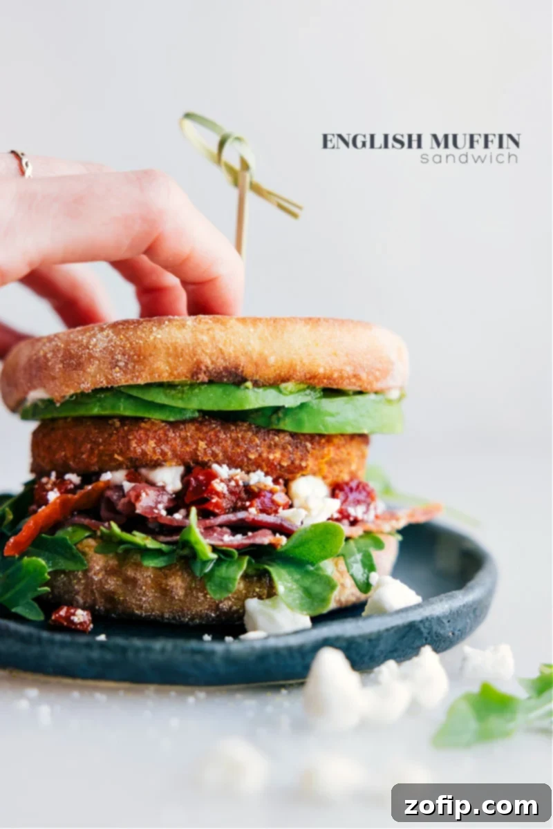 A beautifully assembled English Muffin Sandwich on a plate, showcasing its vibrant layers of avocado, pesto, feta, bacon, and a golden-crisp sweet potato hash brown