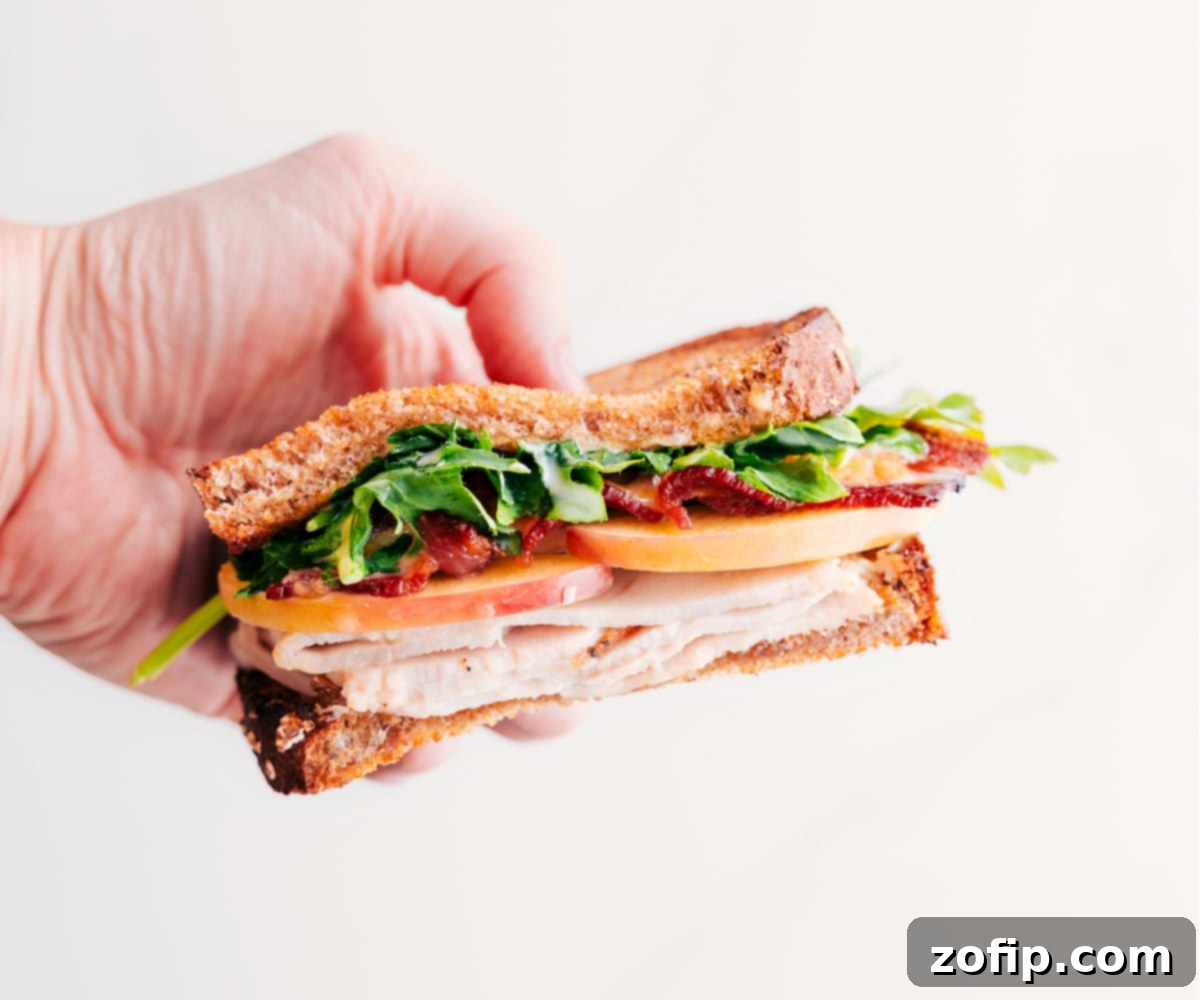A perfectly halved turkey sandwich held up, revealing the delicious layers of turkey, crispy bacon, thinly sliced apple, melted Gouda, and fresh greens, ready to eat.