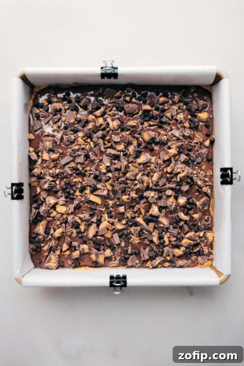 An overhead shot of the finished Pretzel Peanut Butter Bars in a parchment-lined baking pan, sliced and ready to be enjoyed, garnished with chopped peanut butter cups and chocolate chips.