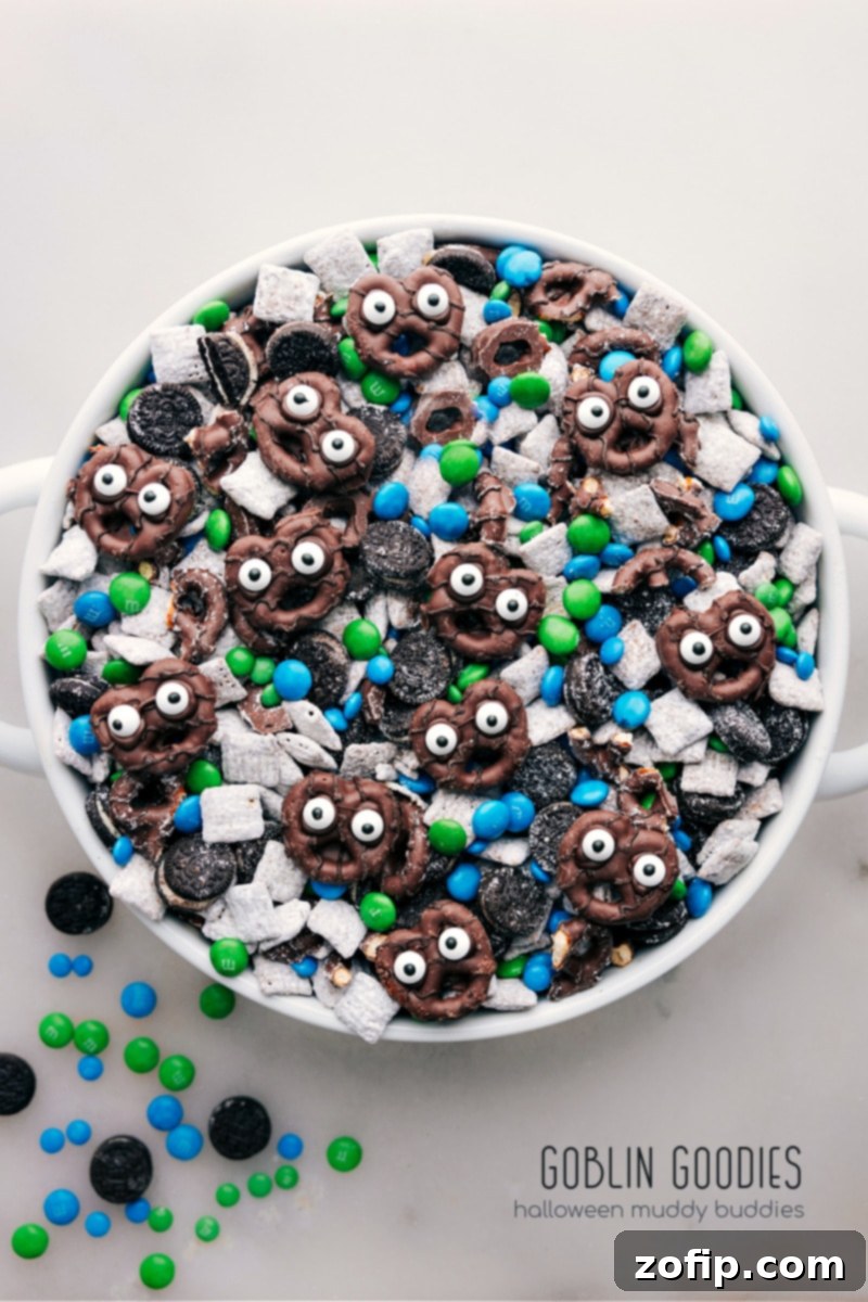 Overhead view of a bowl of Goblin Goodies (Halloween Muddy Buddies) overflowing with colorful M&M's, crushed Oreos, and chocolate-covered pretzels with edible eyes.