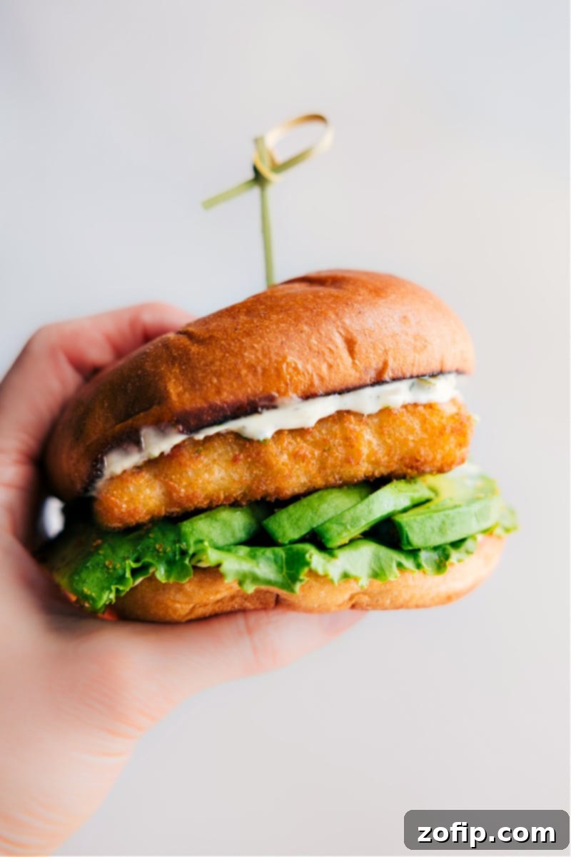 A hand holding a perfectly assembled crispy fish sandwich with golden fish, green lettuce, and creamy avocado, ready for a bite.