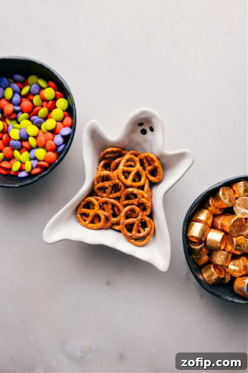 A close-up photograph showcasing the three primary ingredients required for Monster Eyeballs: a bag of vibrant M&M's, a bag of classic pretzel twists, and a roll of individually wrapped Rolo caramel candies.