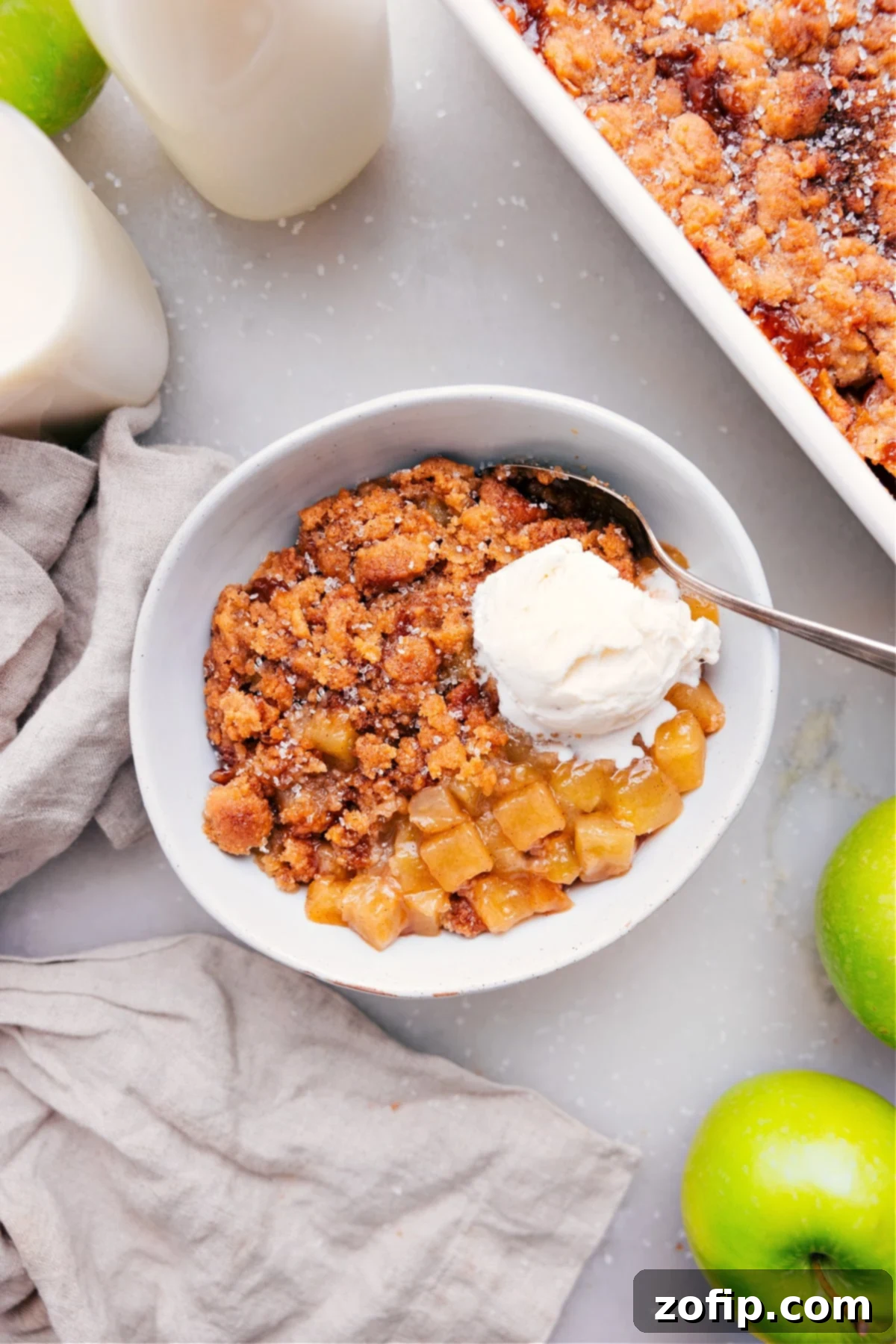 A serving of warm Apple Cobbler topped with a scoop of vanilla ice cream in a white bowl, with a spoon ready to dig in. The cobbler has a golden-brown, crumbly topping and tender apple filling.