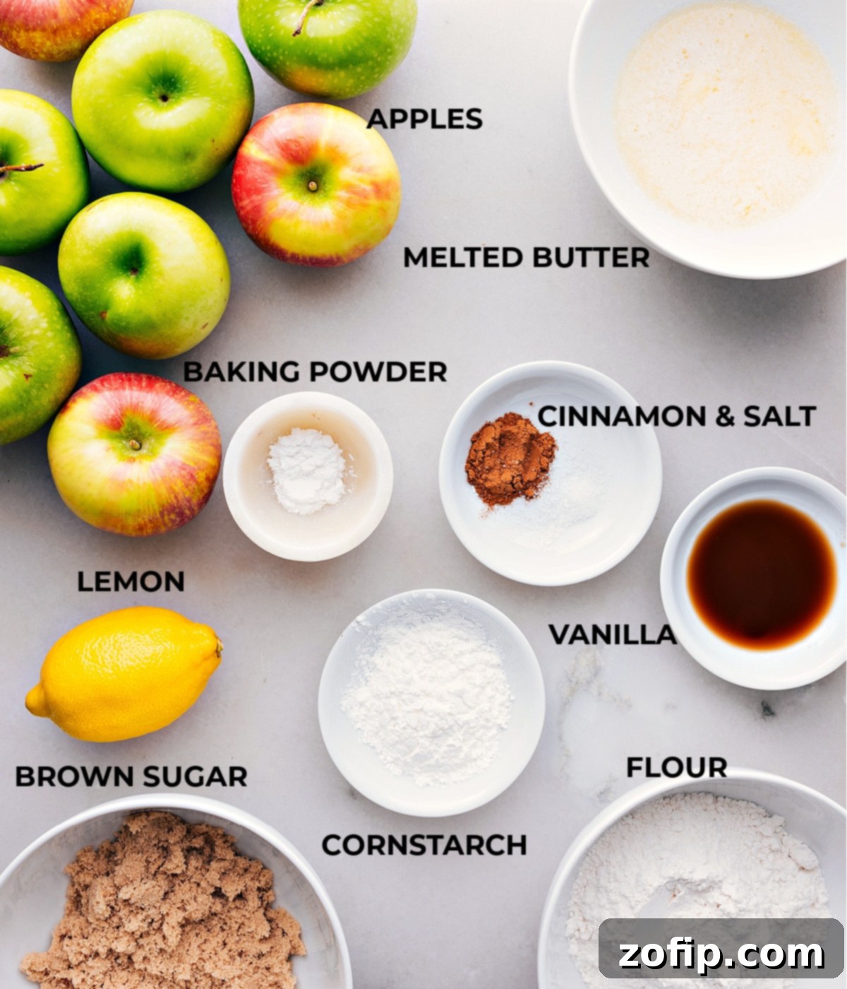 Various ingredients for an apple cobbler recipe laid out on a white surface: fresh apples, melted butter, cinnamon sticks, salt, vanilla extract, baking powder, cornstarch, flour, and brown sugar.