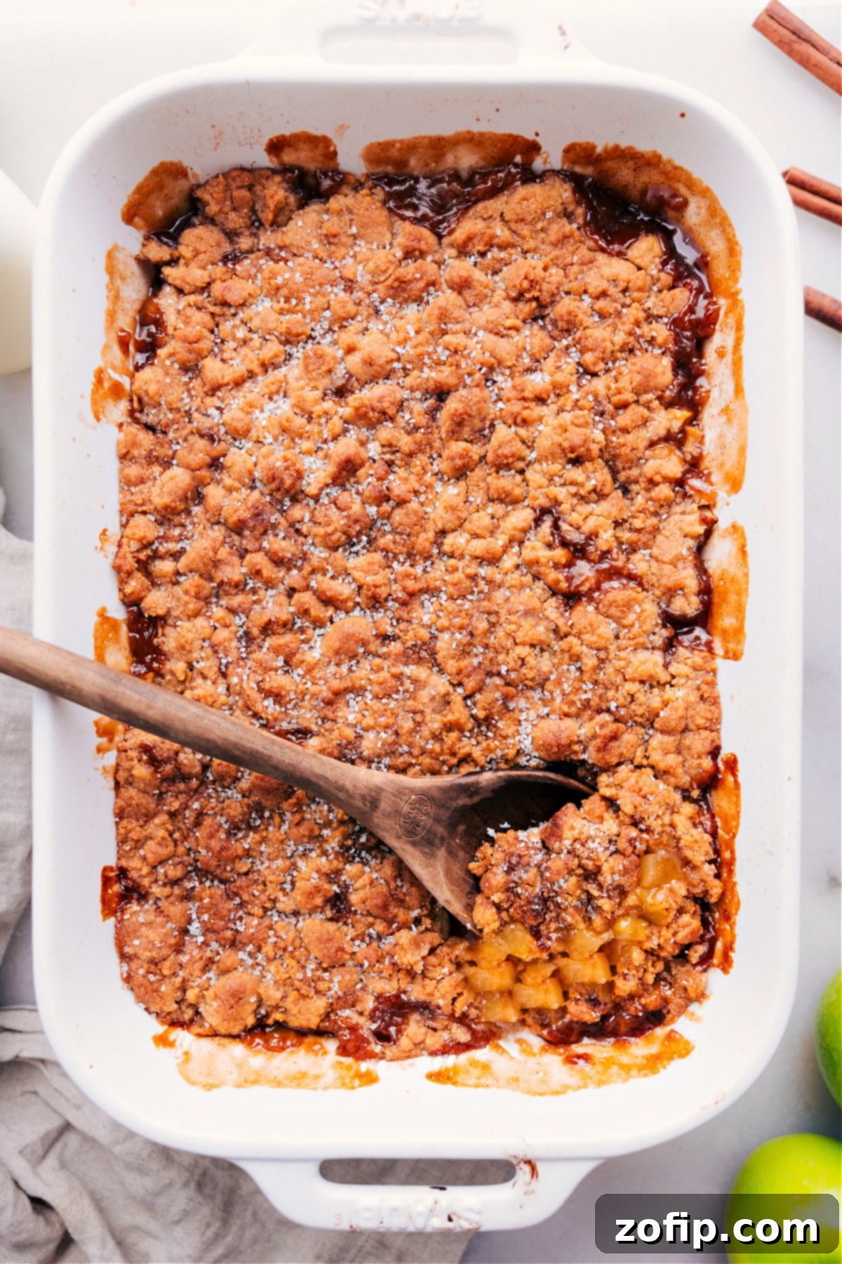 A close-up of a freshly baked apple cobbler pie with a serving spoon already taking a scoop out, showcasing the warm apple filling and crispy topping.