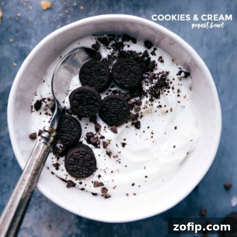 A delightful Cookies and Cream Yogurt Bowl, featuring creamy yogurt topped with crushed miniature Oreos and a mix of chopped dark chocolate and mini chocolate chips, served with a spoon.