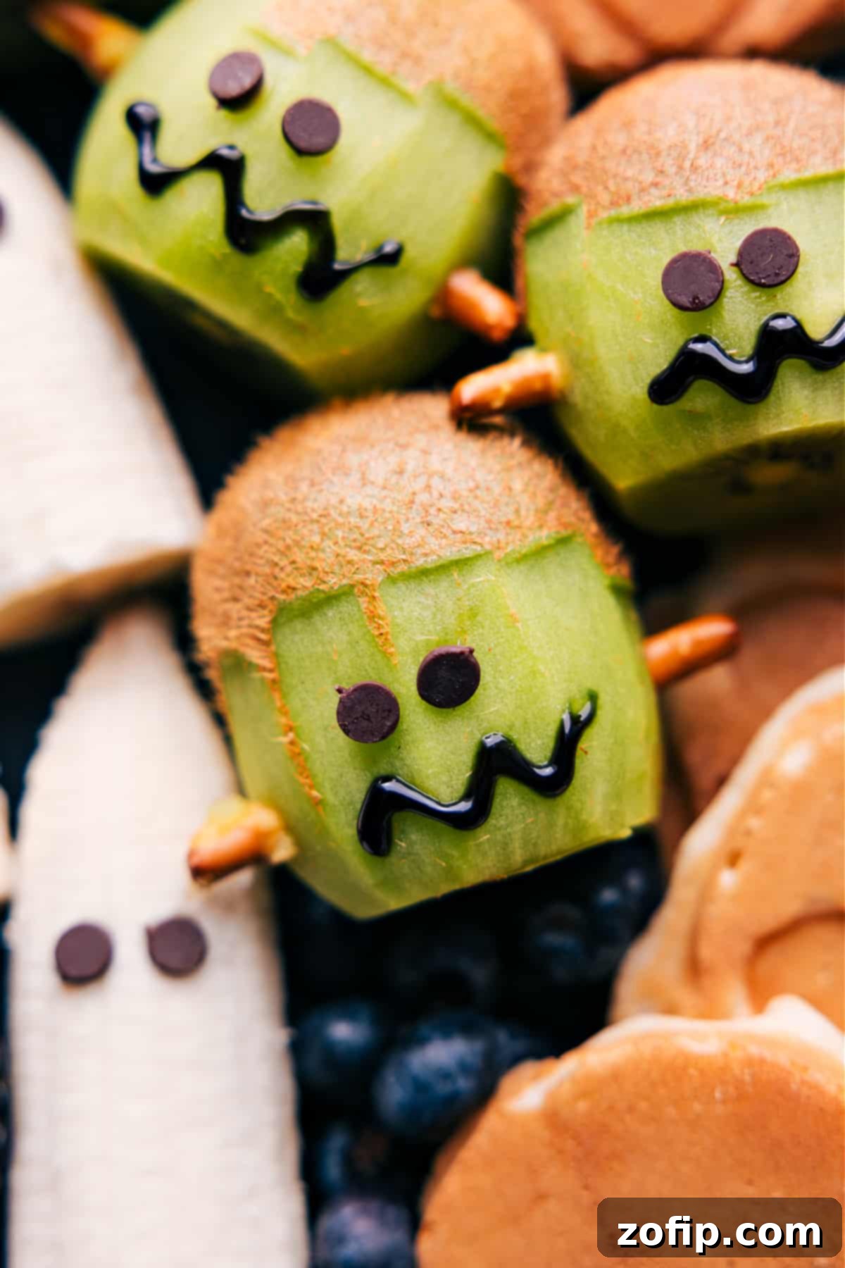 A close-up of a kiwi carved and decorated to resemble Frankenstein's face, complete with pretzel stick 'bolts' and chocolate chip eyes, ready to be served.