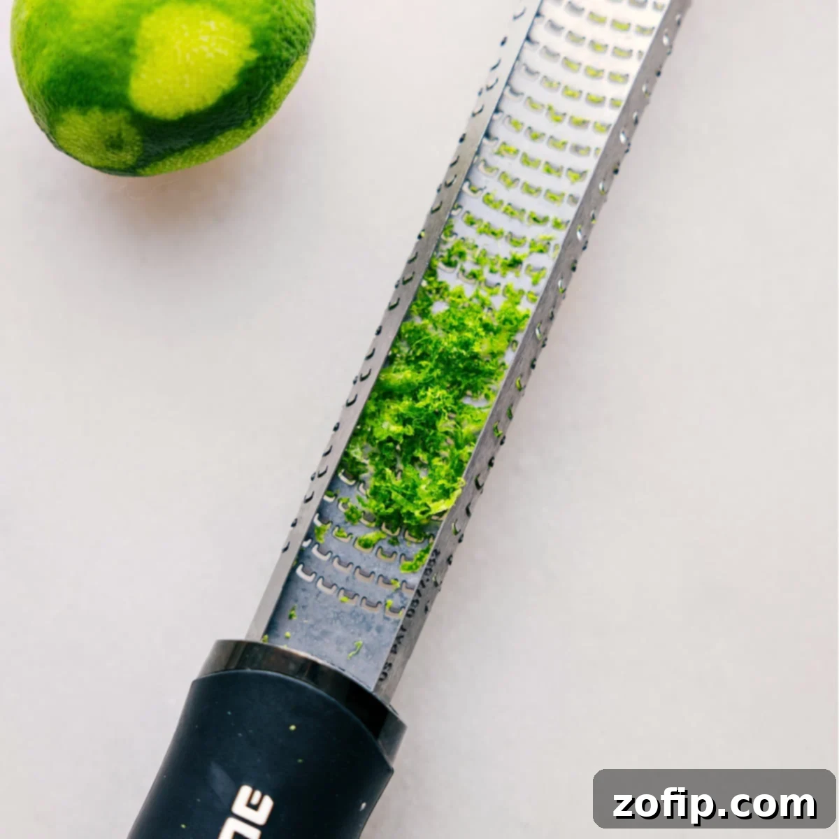A zester tool covered in fine lime peel shavings, highlighting the fresh, green zest just removed from the fruit, ready for use in this recipe.