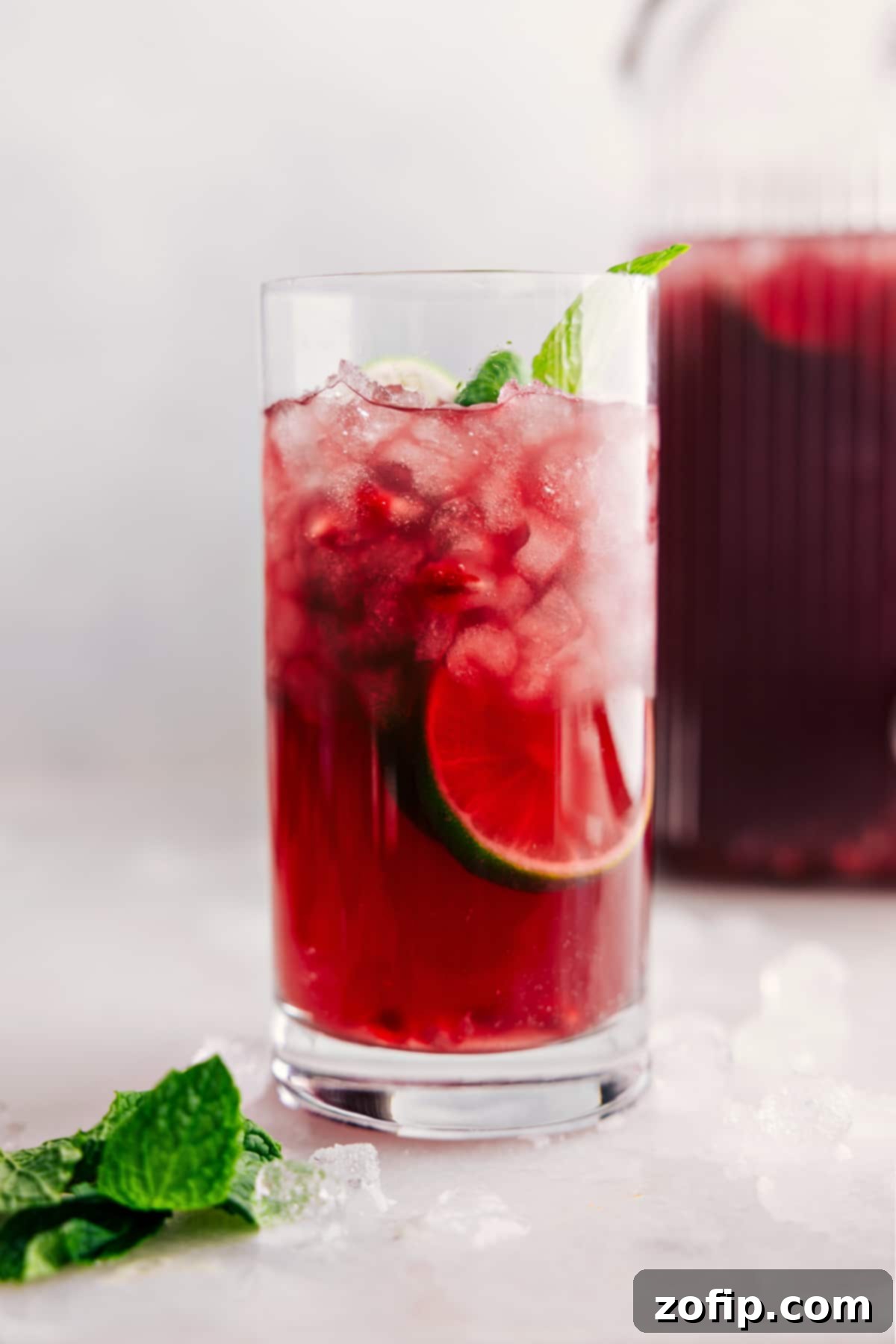 A large glass pitcher filled with vibrant red Christmas punch, garnished with fresh lime slices, pomegranate arils, and mint sprigs, ready to be poured and enjoyed.