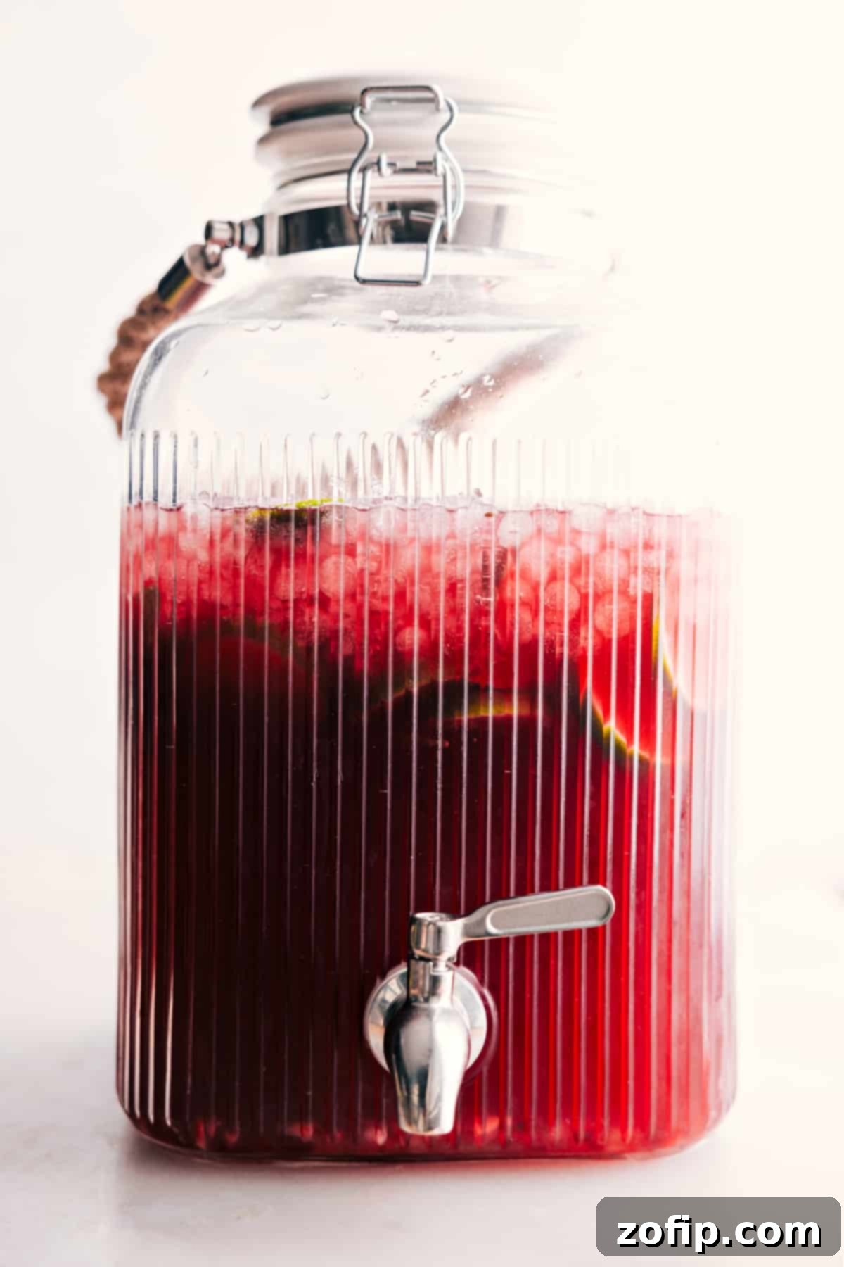 A large glass drink dispenser filled with beautiful red Christmas punch, showcasing lime slices and pomegranate arils, with a spigot ready for serving. It sits on a table alongside a festive setup.