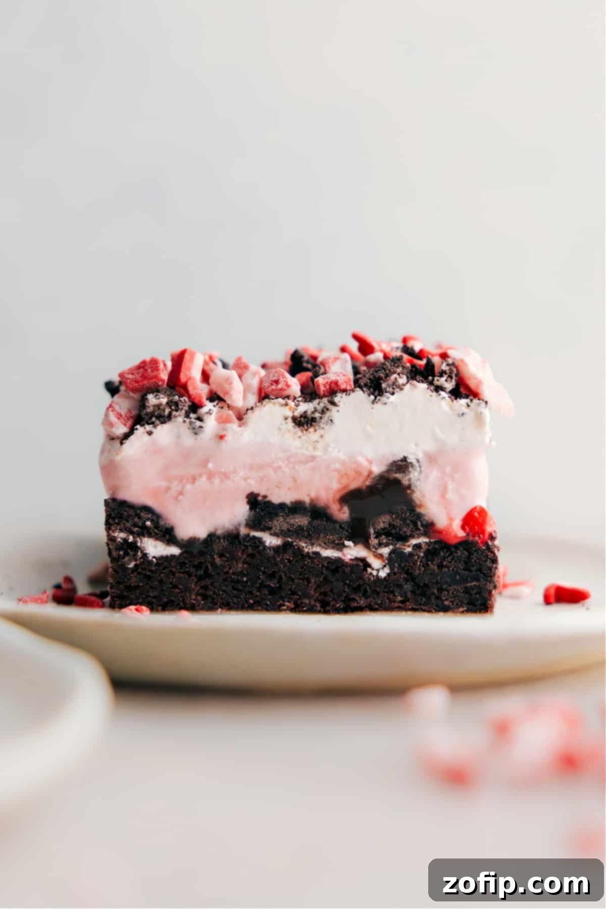 A perfectly sliced Peppermint Brownie Ice Cream Bar showcasing its distinct layers: a dark, dense brownie base, a middle layer of creamy light pink peppermint ice cream, and a generous topping of white whipped cream adorned with vibrant red and green peppermint pieces. The texture appears invitingly rich and cold.