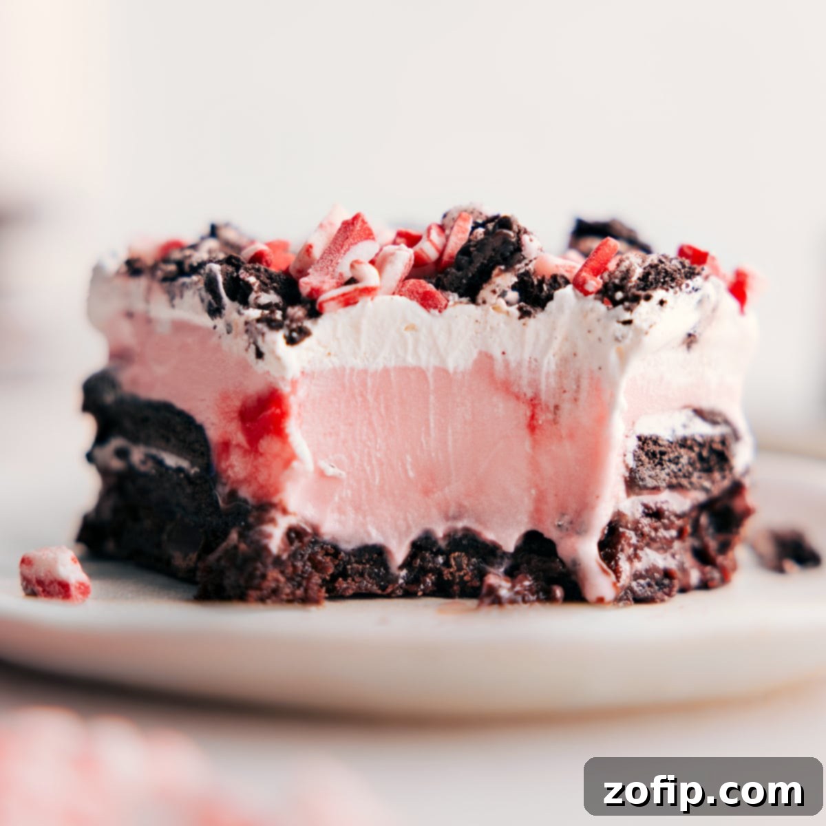 A beautifully presented Peppermint Brownie Ice Cream Bar with a bite taken out, showcasing the distinct layers of brownie, Oreos, fudge, peppermint ice cream, and whipped cream, highlighting the dessert's irresistible combination of flavors and textures.
