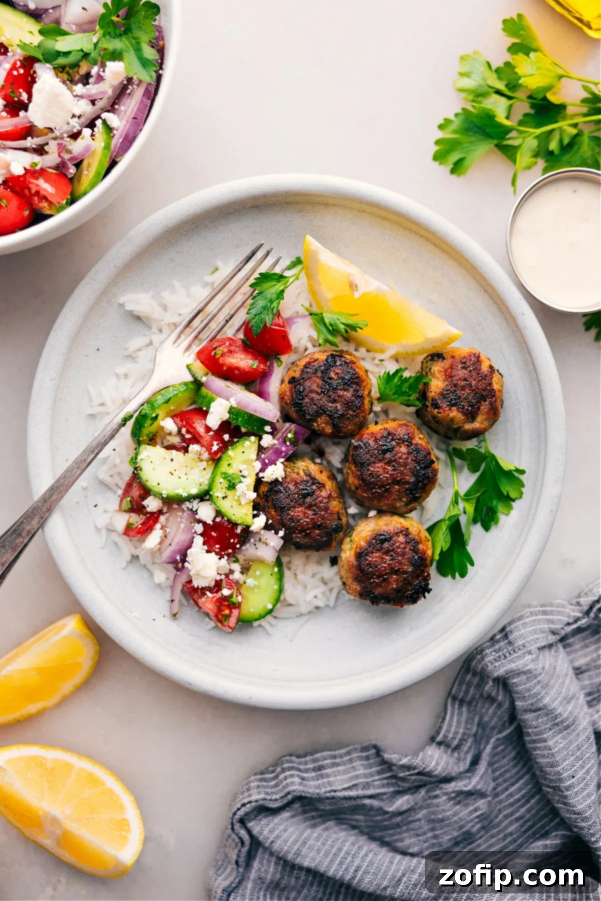 A serving of Greek Turkey Meatballs nestled on a bed of fluffy rice, accompanied by a refreshing cucumber tomato salad, all drizzled with creamy feta sauce.